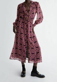 Lång, knapplång klänning i ett djärvt rosa och lila geometriskt mönster. Har en krage, elastisk midja och genomskinliga långa ärmar. Bärs med glänsande svarta loafers.