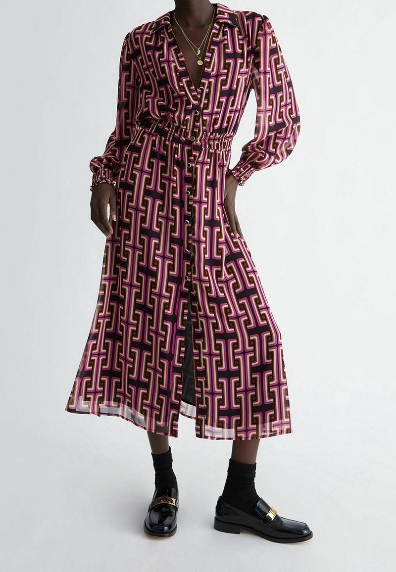 Lång, knapplång klänning i ett djärvt rosa och lila geometriskt mönster. Har en krage, elastisk midja och genomskinliga långa ärmar. Bärs med glänsande svarta loafers.