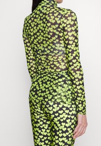 Langarmoberteil und enganliegende Leggings aus transparenter schwarzer Stoff mit kleinen, leuchtend grünen Herzen, ausgestattet mit einem Stehkragen und einem körperbetonten Design.