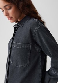 Frau mit geschlossenen Augen, die ein dunkelgraues Jeanshemd mit einer Brusttasche trägt, steht vor einem schlichten hellen Hintergrund.