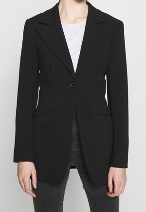 Personne portant un blazer noir cintré avec un bouton, associé à un top blanc et un jean gris foncé, debout devant un fond uni.