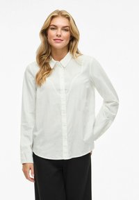 Camicia bianca a maniche lunghe con bottoni, presenta un colletto classico, tessuto morbido e un orlo leggermente curvato. Indossata con pantaloni neri.