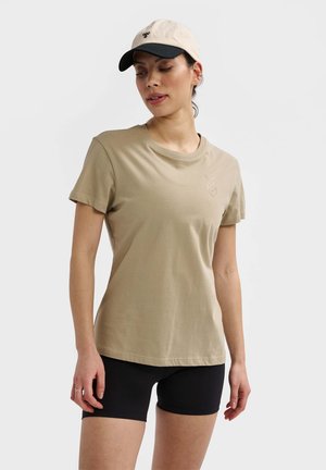 Beige bomulds t-shirt med korte ærmer, rund halsudskæring og subtile broderede chevron-detaljer. Båret med sorte træningsshorts og kasket.