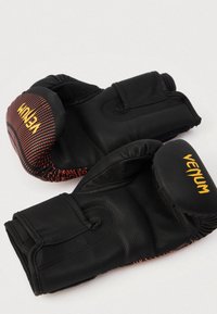 Μαύρα γάντια πυγμαχίας με κόκκινες διακοσμητικές λεπτομέρειες. Κατασκευασμένα από συνθετικό υλικό, με ιμάντες βέλκρο και ανάγλυφα λογότυπα μάρκας στην εξωτερική πλευρά.