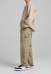 Beige huppari, jossa on rosoiset olkapäät, yhdistettynä vaalean khaki värisiin cargo-housuihin, joissa on sivutaskut. Mustat, punaiset ja valkoiset tennarit täydentävät asua.