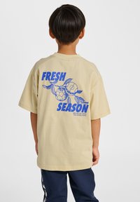 Beiges T-Shirt aus Baumwolle mit kurzen Ärmeln, das auf der Rückseite ein blaues grafisches Design mit dem Text "FRESH SEASON" und fußballbezogenen Elementen zeigt.