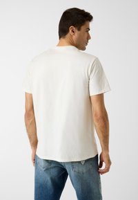 T-shirt à manches courtes de couleur crème claire avec un col rond, fabriqué en coton doux, porté avec un jean bleu délavé. Design simple et minimaliste.