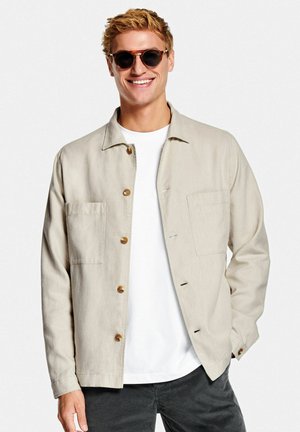 Beige Button-Up-Jacke aus leichtem Stoff, mit zwei vorderen Taschen, einem spitzen Kragen und braunen Knöpfen. Getragen über einem weißen Hemd.