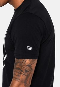 Musta puuvillainen t-paita, jossa on lyhyet hihat ja pyöreä kaula-aukko. Vasemman hihan päällä on valkoinen brodeerattu logo.