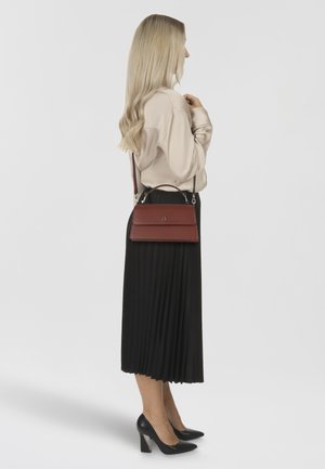 Borsa a mano in pelle rossa con patta anteriore piatta, tracolla regolabile e accessori in argento. Indossata con una blusa color crema e una gonna plissettata nera.