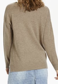 Beżowy sweter dziany o luźnym kroju, z prążkowanym dołem i długimi rękawami; noszony na jasnoniebieskich spodniach dżinsowych z wzorem.