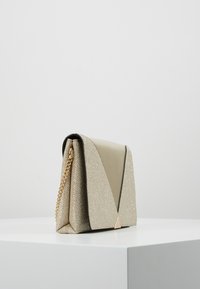 Beżowa i błyszcząca beżowa torebka o strukturze, z złotym łańcuszkiem na ramię, trójkątnymi akcentami oraz klapą złożoną na zakładkę. Teksturowane powierzchnie.