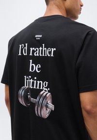 Άνδρας φορεί μαύρο t-shirt Adidas με το κείμενο «Προτιμώ να σηκώνω βάρη.» και γραφικό αλτήρα στην πλάτη, γυρισμένος ελαφρώς στο πλάι.