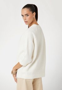 Pull en tricot crème surdimensionné avec des manches courtes, présentant une texture côtelée et une couture décorative le long de l'ourlet. Design court à l'arrière.