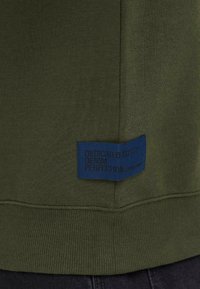 Olivengrønn strikket stoff med marineblå rektangulær etikett som leser "DEDICATED TO DENIM PERFECTION" sydd nær sømmen.