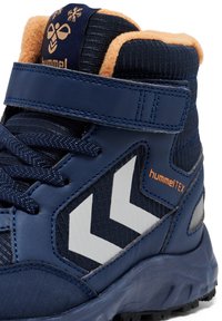 Bota de senderismo en color navy con un upper de cuero suave, acentos de textil, correa de Velcro, detalle en chevrón blanco y logotipo hummelTEX en naranja.