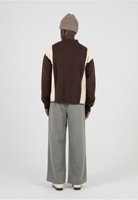 Pullover marrone e crema con motivo, colletto alto, abbinato a pantaloni grigi larghi e un berretto di lana beige. Tessuto morbido, vestibilità casual.