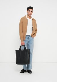 Fekete bőr válltáska lapos fogantyúkkal, függőleges varráselemekkel. A modellen bézs kardigán, fehér ing és világoskék farmer van.