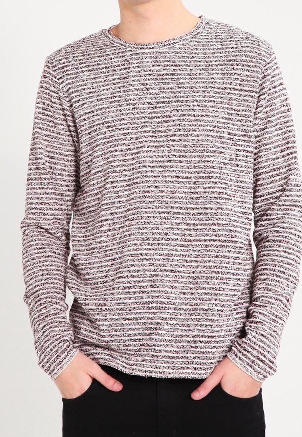 Homme portant un pull à manches longues et col rond avec des rayures horizontales noires et blanches, les mains dans les poches, devant un fond uni.