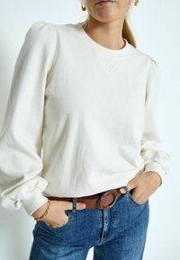 Felpa bianca con maniche a palloncino, scollatura rotonda e tessuto strutturato. Indossata con jeans blu scuro e una cintura marrone con fibbia circolare.