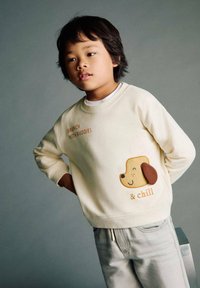 Sweatshirt crema con stampa "BRUNCH WITH BUDDIES" e applicazione di un cane marrone, abbinato a pantaloni grigi chiari. Tessuto morbido e vestibilità rilassata.