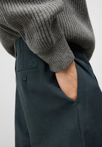 Maglione grigio a coste sovrapposto a pantaloni verde scuro con una tasca laterale e una tasca posteriore con bottone e logo ricamato.