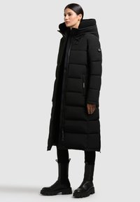 Cămașă puffer neagră cu un design lung, matlasat, glugă mare, buzunare laterale și o textură netedă. Purtați-o peste pantaloni negri și cizme.