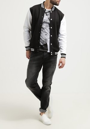 Men's black and white varsity jacket with striped cuffs, buttons, and a gray graphic t-shirt underneath, paired with dark jeans and white slip-on shoes.