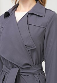 Szary płaszcz typu wrap z dużym kołnierzem, wyposażony w pasek w talii i gładką tkaninę. Widoczne są szczegółowe przeszycia oraz boczne kieszenie.