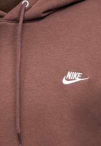 Rjava jopa iz mehkega blaga z žepom spredaj, z belo Nike značko na prsih in nastavljivo kapuco z vrvico.