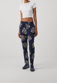 Pantalon bleu marine à imprimé floral avec des motifs de roses blanches, taille élastique et cordelette. Associé à un t-shirt blanc court et des baskets bleues.