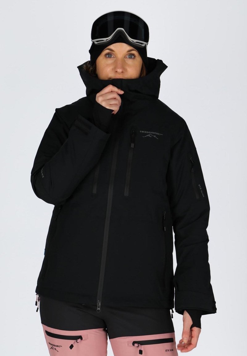 Black snow jacket with multiple zippered pockets, high collar, adjustable cuffs, and ventilation zips, paired with pink ski pants.