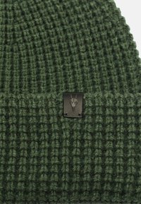 Green knitted beanie with a ribbed texture and a black metal tag featuring a ram skull design. Quality wool blend material.