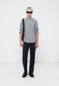 Grau Hemd mit Knöpfen, dunkle Hose, schwarze Sonnenbrille und Schuhe. Slim-Fit, lange Ärmel, einzelne Brusttasche, glatter Stoff, minimalistisches Design.