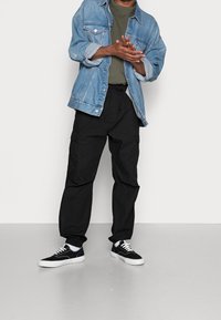 Leren spijkerjacket in lichtblauw met twee borstzakken, olijfgroen T-shirt, zwarte cargo broek met zijzakken en zwarte sneakers met witte zolen.