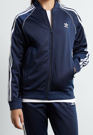 Person iført navyblå Adidas træningsjakke og matchende bukser med hvide striber på ærmer og ben, stående med den ene hånd i lommen.