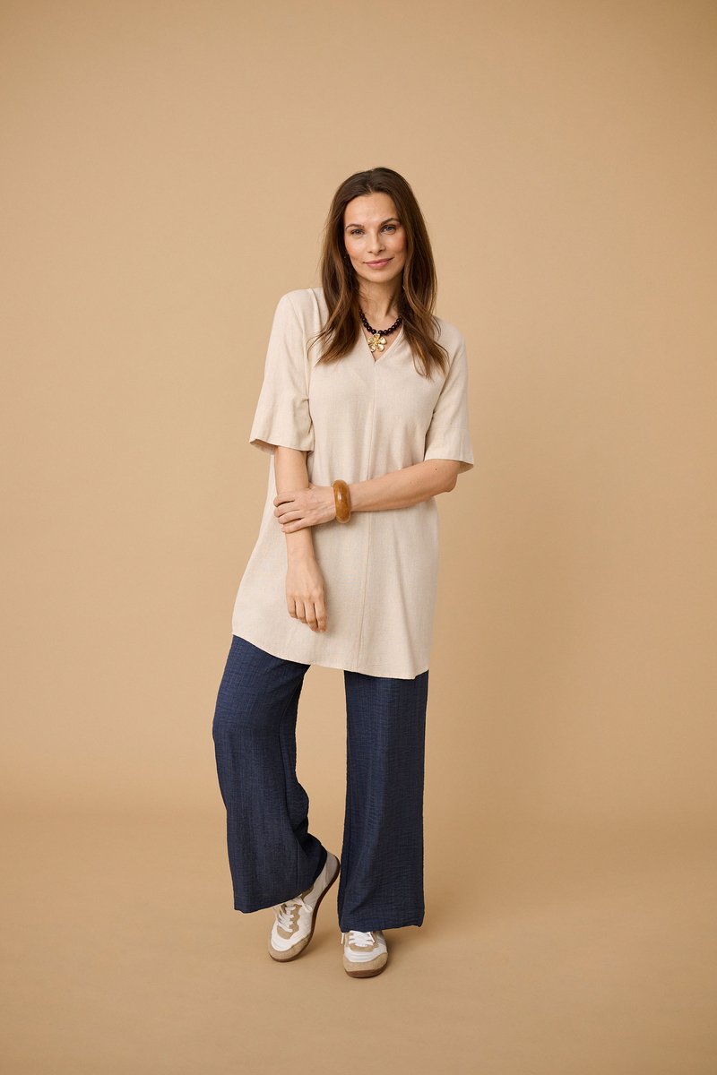 Vrouw poseert tegen een beige achtergrond, draagt een beige tuniek, marineblauwe wijde broek, witte sneakers, een stevige armband en gelaagde kettingen.