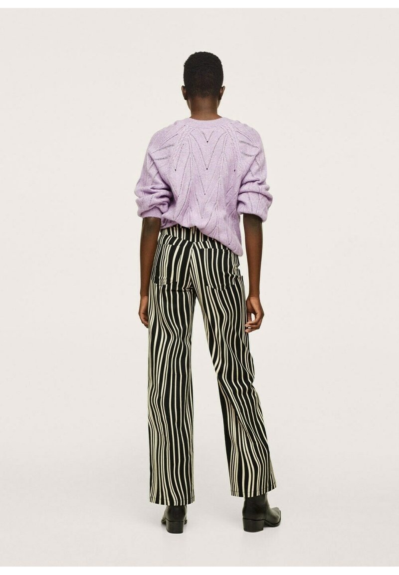 Personne portant un pull tricoté violet clair et un pantalon à rayures verticales noir et blanc, se tenant de dos sur un fond uni.