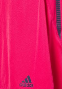 Camisa deportiva rosa hecha de una tela suave, con un logo de Adidas en azul oscuro y un acento gris con patrón a lo largo de la costura lateral.