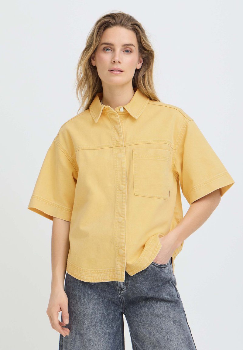 Femme aux cheveux châtain clair portant une chemise jaune à manches courtes boutonnée et un jean noir délavé, debout avec une main dans la poche.