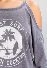 Sweat-shirt gris à épaules dénudées avec un motif estompé de palmier et planche de surf, doté d'une coupe décontractée et d'un tissu doux et texturé.