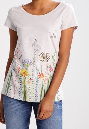 Femme portant une chemise blanche à manches courtes avec des motifs colorés de fleurs sauvages et de papillons, associée à un jean bleu.