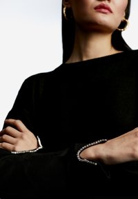 Černý svetr s dlouhými rukávy, s stříbrným korálkovým lemem na manžetách. Model má zkřížené ruce, což ukazuje na hladkou texturu látky.