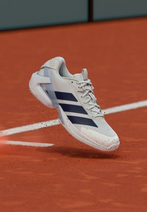 Scarpa da tennis bianca con strisce blu navy, tomaia in mesh sintetico, suola testurizzata e dettagli traslucidi, mostrata in aria sopra un campo in terra rossa.