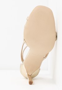 Metallic gold high-heeled sandal with a sleek pointed toe, strappy upper, and a textured light tan sole. Features a slender heel.