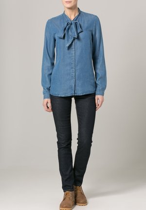 Blouse en denim bleu clair avec un nœud au col, manches longues et une coupe ajustée, associée à un jean foncé et des bottes marron.