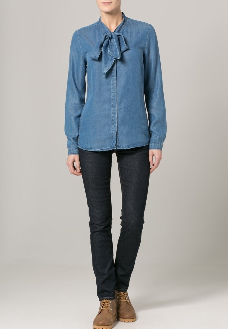 Blusa de mezclilla azul clara con un lazo en el cuello, de mangas largas y un diseño ajustado, combinada con jeans oscuros y botas marrones.