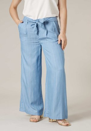 Femme portant un pantalon large bleu clair avec ceinture nouée, haut blanc et sandales à talon bloc doré et ouvert aux orteils, debout, les mains le long du corps.