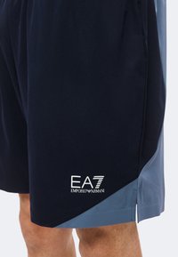 Ναυτικό και μπλε σορτς με το λογότυπο Emporio Armani EA7 στο αριστερό πόδι, που φοριούνται από ένα άτομο που δείχνει την περιοχή του μηρού και του γόνατου.