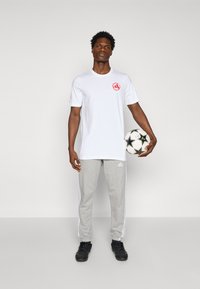 White short-sleeve T-shirt with red logo, gray sweatpants with white stripes, and black athletic shoes. Holding a black and white soccer ball.